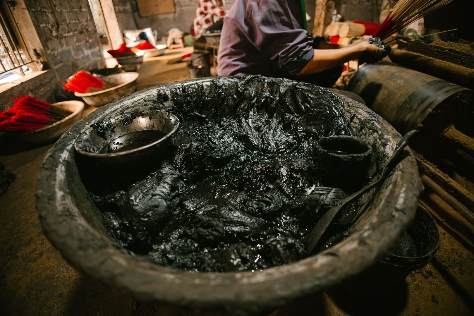 Large vat of black dye beside bundles of red fiber in a workshop